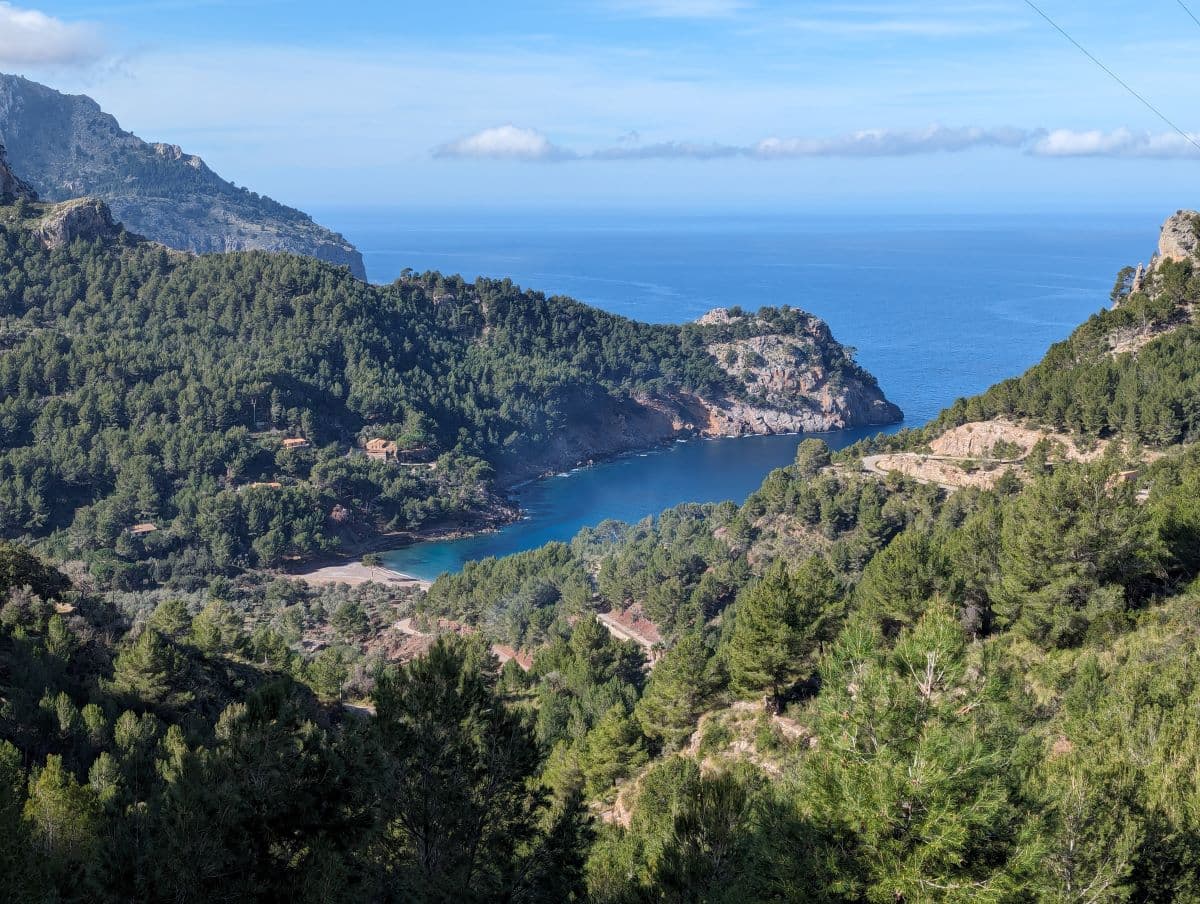 Panoramablick auf Cala Tuent, Mallorca, mit einer abgelegenen Bucht inmitten grüner Hügel, einem Kieselstrand und dem Mittelmeer am Horizont.