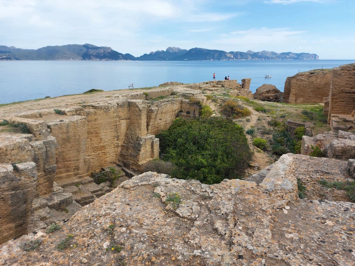 Overlooking ancient stone quarry remains, with a panoramic view of the sea and distant Tramuntana mountains. Linda and other hikers explore the historic site, immersed in the vast, open landscape under a cloudy sky.