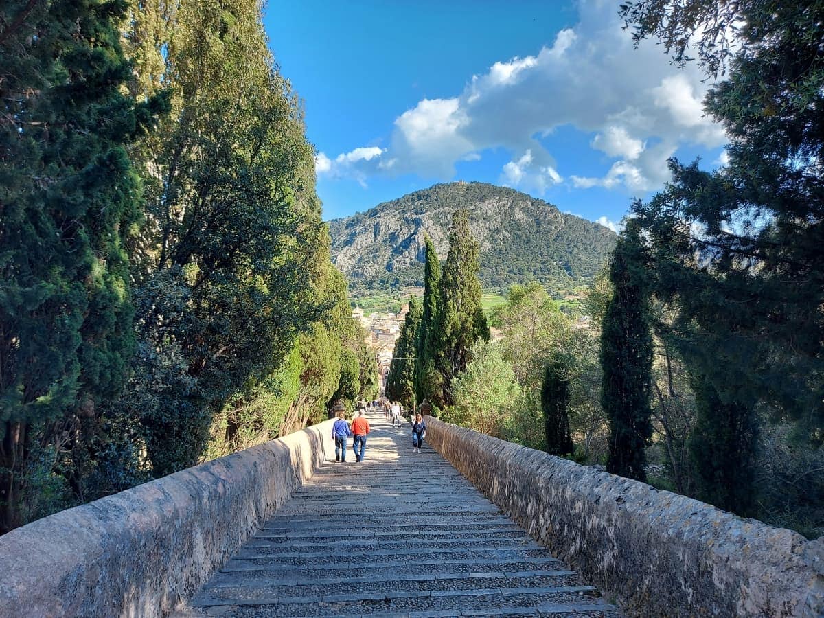 Linda and other tourists walk down the old stone steps of the Calvary in Pollensa Mallorca, surrounded by lush greenery, with a view of a mountain in the background under a partly cloudy sky.