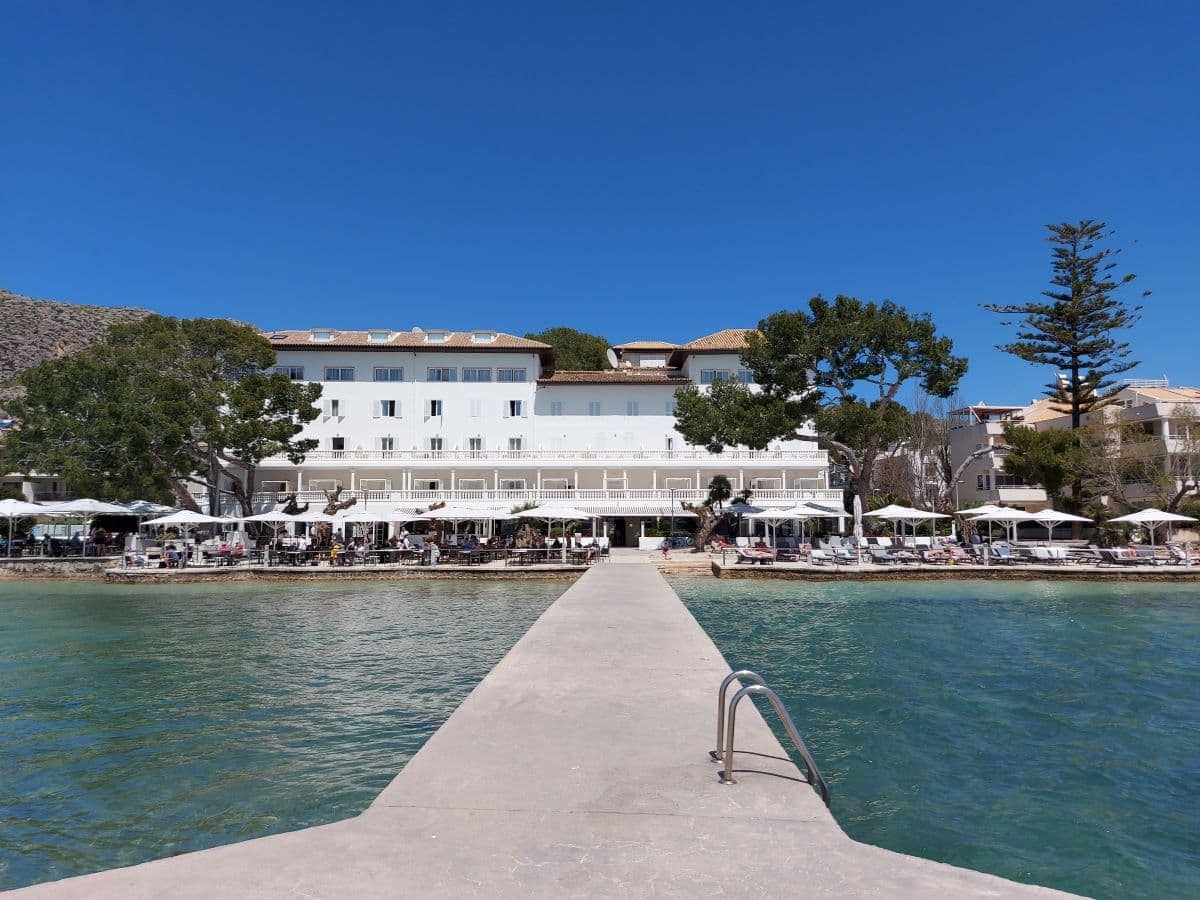 The elegant façade of the Hotel Illa d'Or in Port de Pollença, set against a bright blue sky. It overlooks a concrete jetty that extends into the calm turquoise waters, while guests enjoy the seaside atmosphere under white parasols.