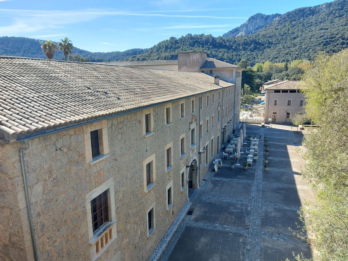 Elevated view of the hotel accommodations at Lluc Monastery, with a series of windows lining the stone building's facade overlooking an outdoor café area, set against the backdrop of the scenic Tramuntana Mountains under a clear sky.