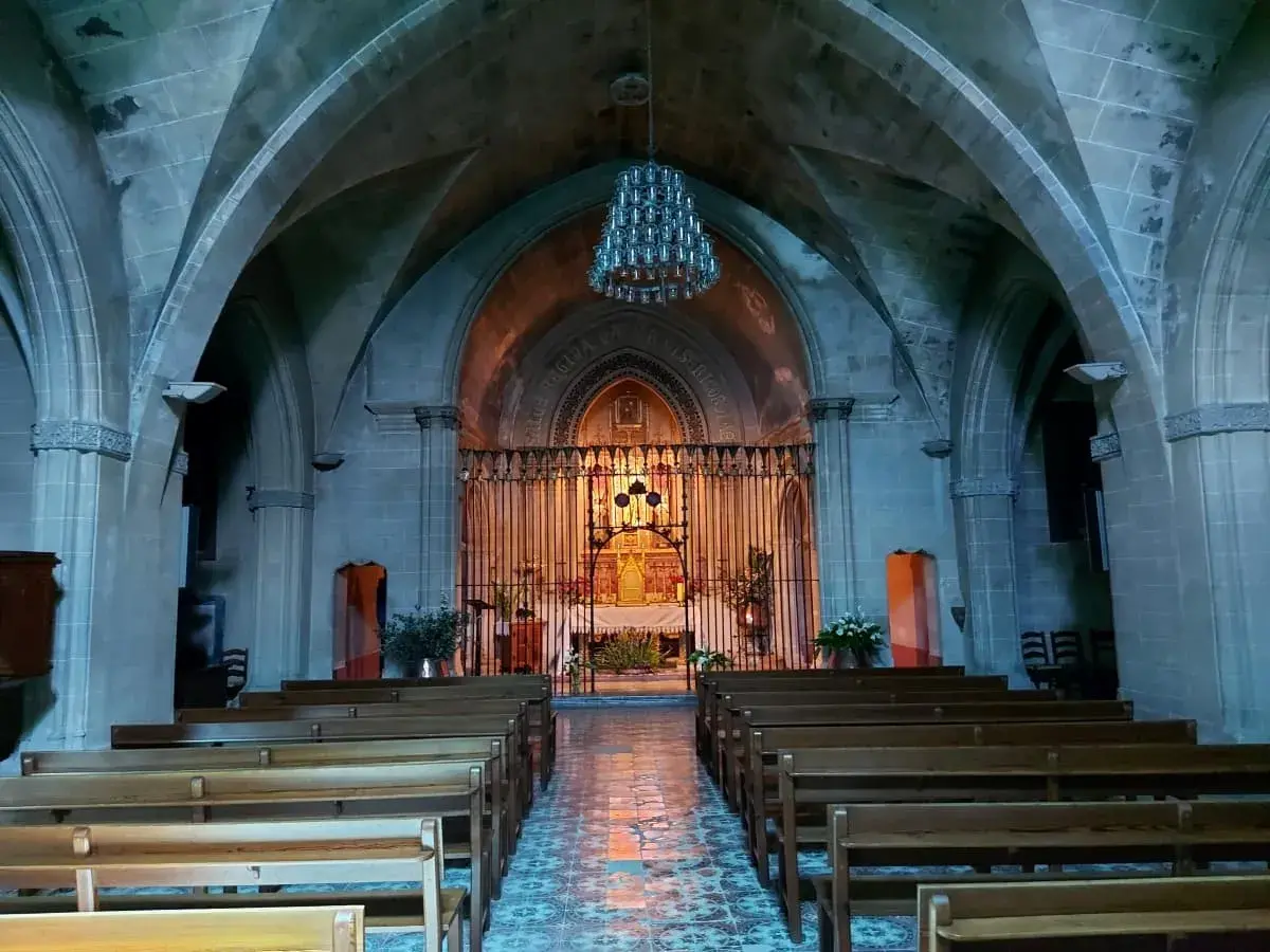The interior of the monastery church on Puig de Maria, featuring rows of wooden pews leading to an ornate altar, with gothic arches overhead and a patterned blue floor, all bathed in soft, ambient lighting.