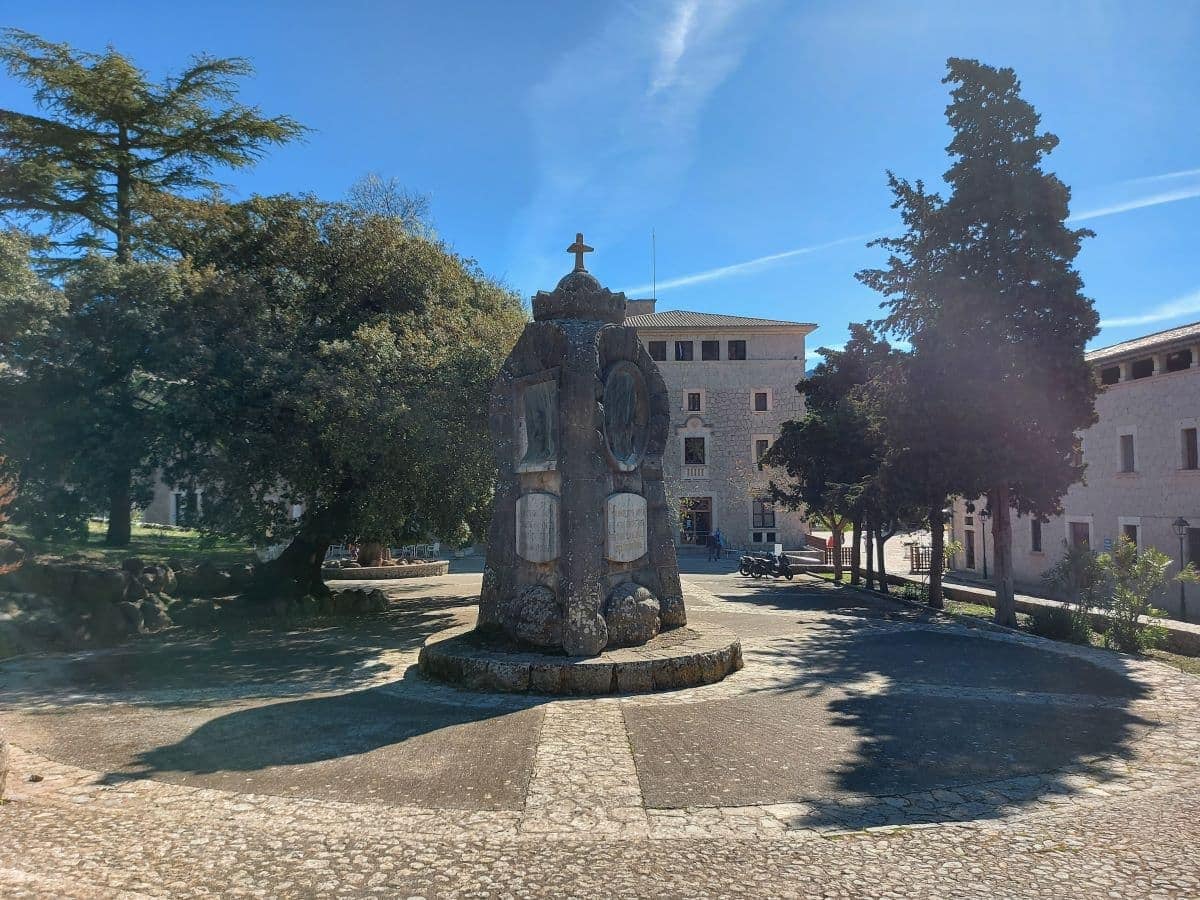 Central view of a rugged stone monument at the Lluc Monastery courtyard with carved religious symbols, set against the monastery buildings and tall trees under the soft light of a sun-drenched sky.