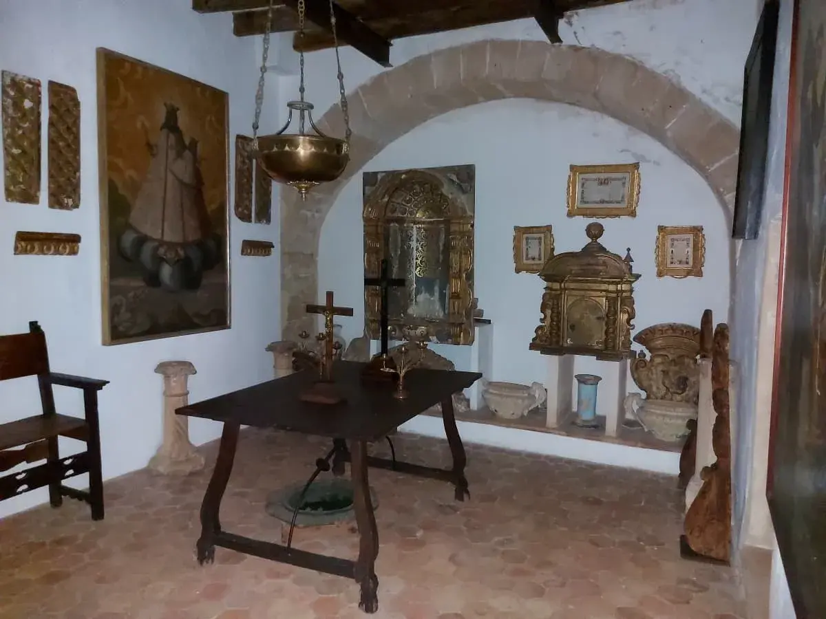 The interior of a room in the Puig de Maria Monastery museum, displaying religious artifacts, a large painting of a saint, ornate golden chests, and antique furniture, under warm lighting.