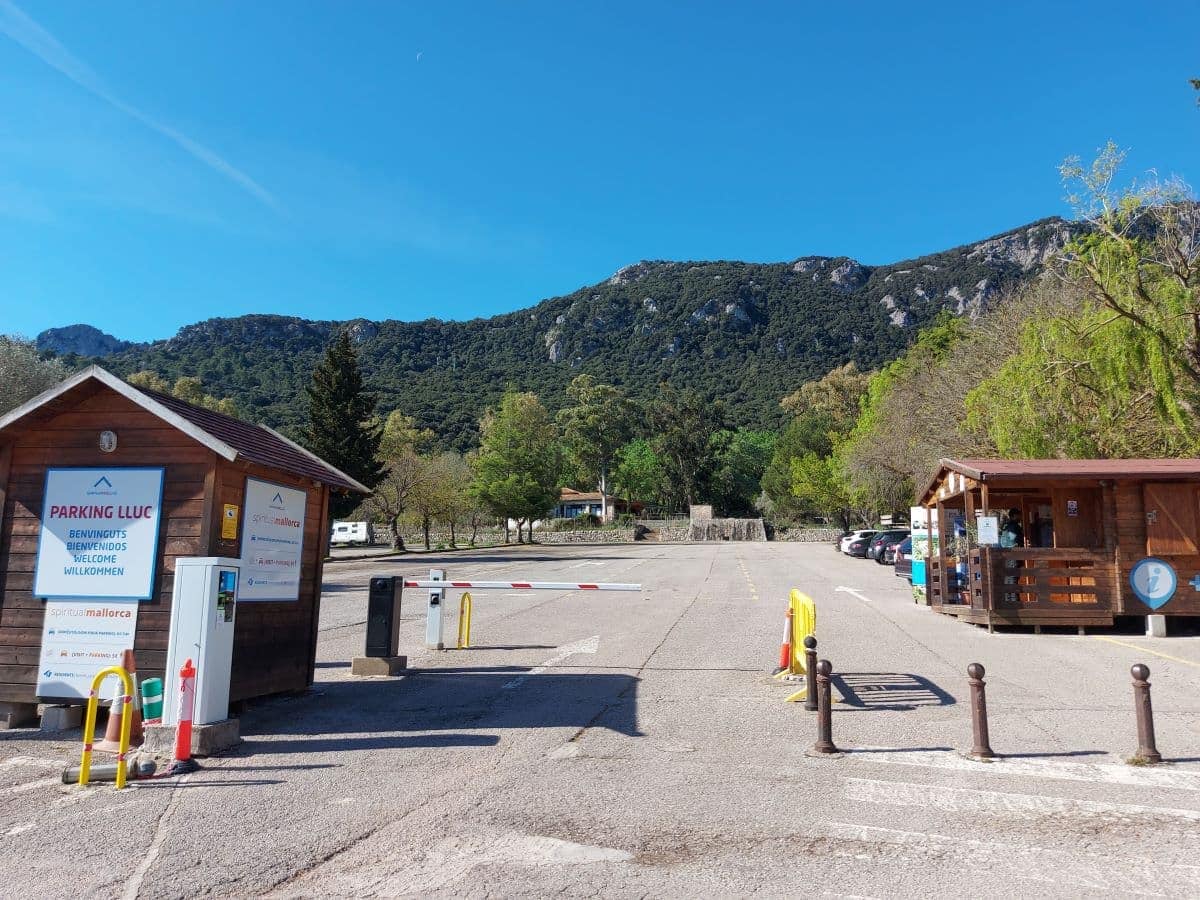 The parking area of Lluc Monastery with clear blue skies above, featuring a welcome sign in multiple languages, a small wooden booth, and a refreshment kiosk, all set against the backdrop of the verdant Tramuntana mountain range.