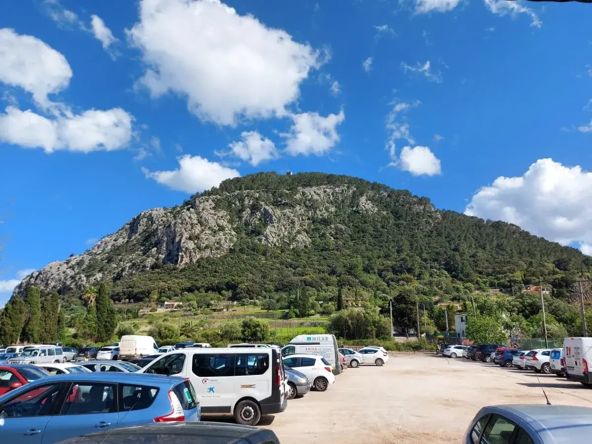 A parking lot at the foot of Puig de Maria, filled with cars, is a popular starting point for visitors who want to explore the mountain. In the background, the impressive wooded mountainside rises under a sky of white clouds.
