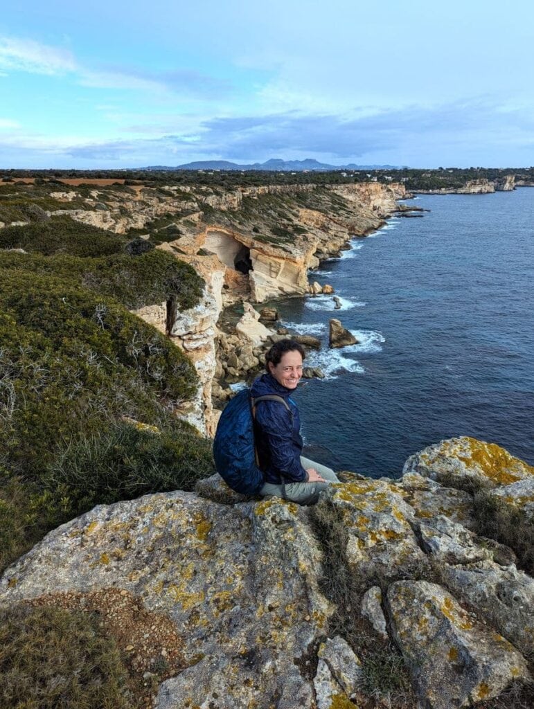 Linda sits on a rugged cliffside, smiling as they overlook the south coast of Mallorca with its dramatic cliffs and the Mediterranean Sea. The overcast sky casts a soft light on the undulating landscape, highlighting the natural beauty of the coastal terrain