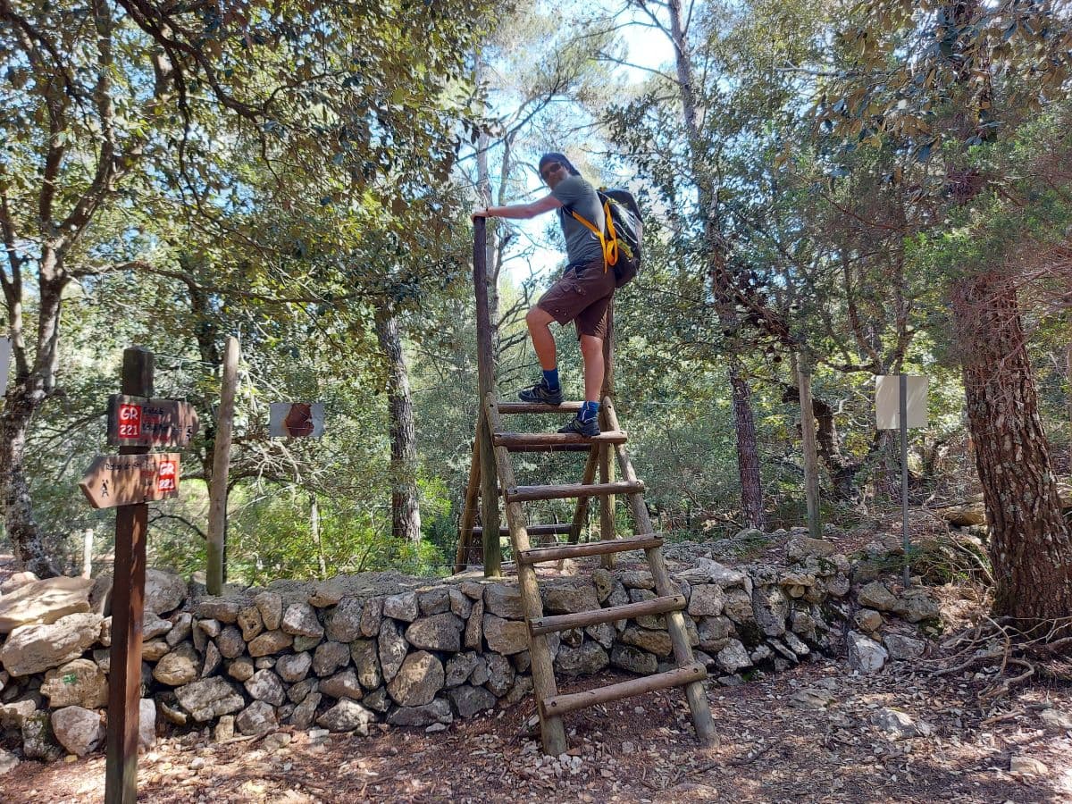 Jan crosses a dry stone wall on a narrow wooden ladder along a hiking trail around Lluc Monastery
