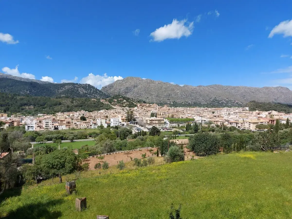 A breathtaking view of Pollença, with its clusters of white and beige buildings nestled among green fields and framed by the majestic Tramuntana mountains under a clear blue sky with scattered clouds.