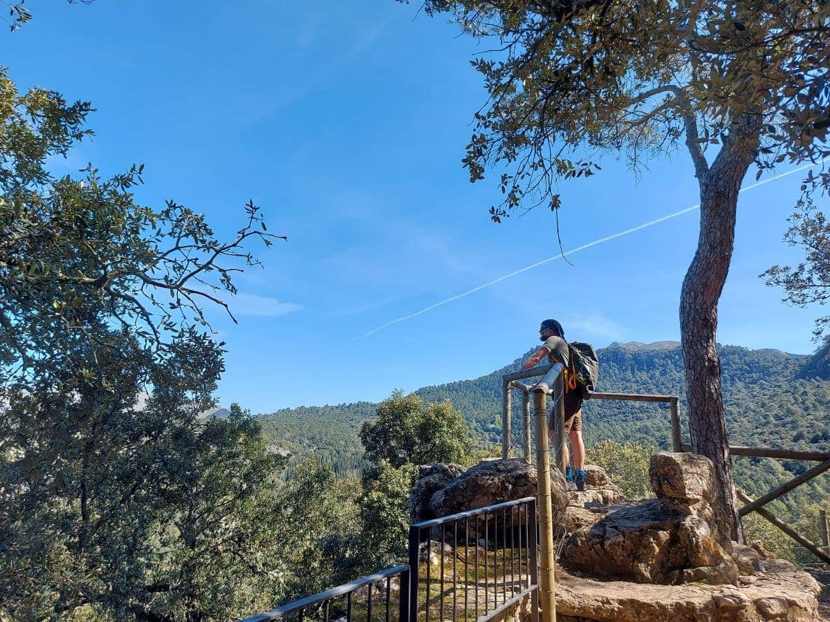 Jan stands at a viewpoint railing at Son Amar, Mallorca, gazing out over the scenic landscape of rolling forested hills under a clear blue sky, with contrails marking the vast expanse above.