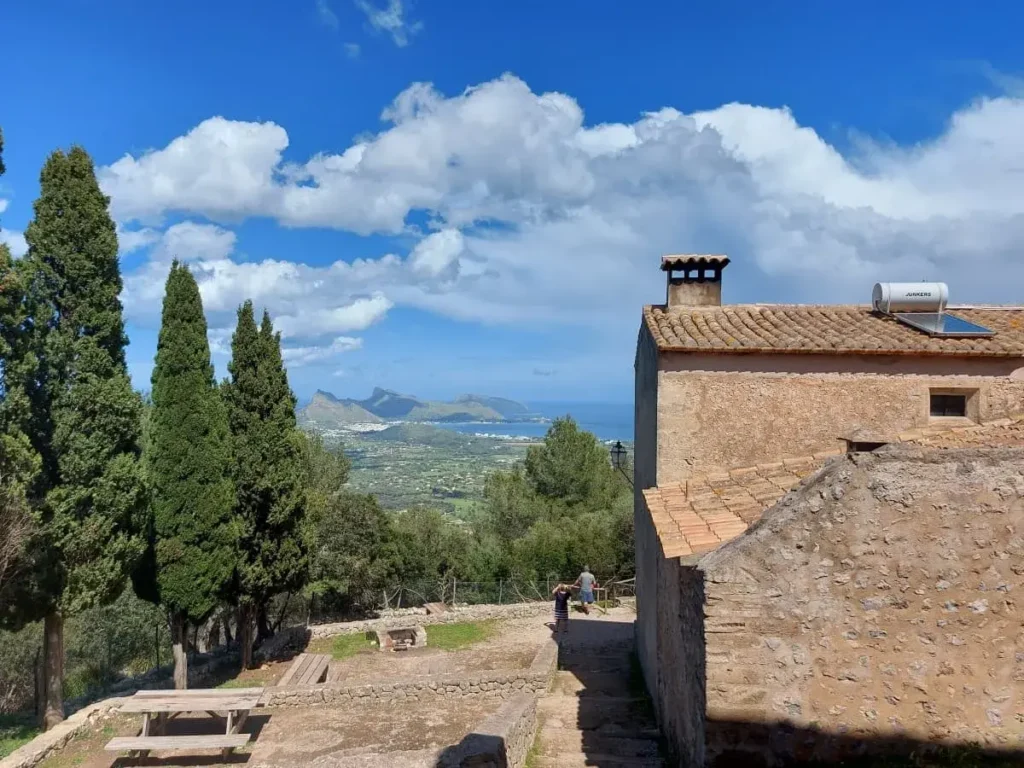 The scenic overlook from Puig de Maria, capturing the contrast of an old stone building with a terracotta roof against the backdrop of a verdant valley, with a clear view to the bay and the distinctive peninsula of Formentor under a sky dotted with fluffy clouds.