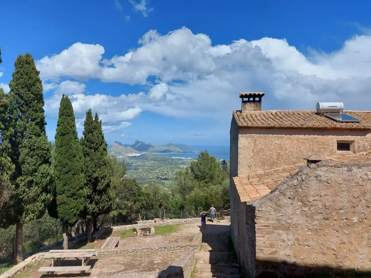 Der Panoramablick vom Puig de Maria, der den Kontrast eines alten Steingebäudes mit Terrakotta-Dach vor dem Hintergrund eines grünen Tals einfängt, mit freiem Blick auf die Bucht und die markante Halbinsel Formentor unter einem Himmel, der mit flauschigen Wolken übersät ist.