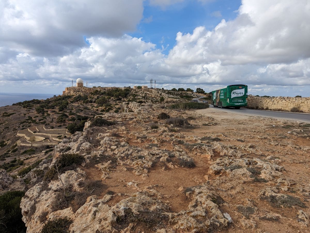 Ein grüner öffentlicher Bus mit der Aufschrift „Knorr“ fährt entlang einer Straße neben den schroffen Dingli-Klippen auf Malta. Die natürliche Kalksteinlandschaft unter einem teilweise bewölkten Himmel bildet eine ruhige Kulisse für die Busroute.