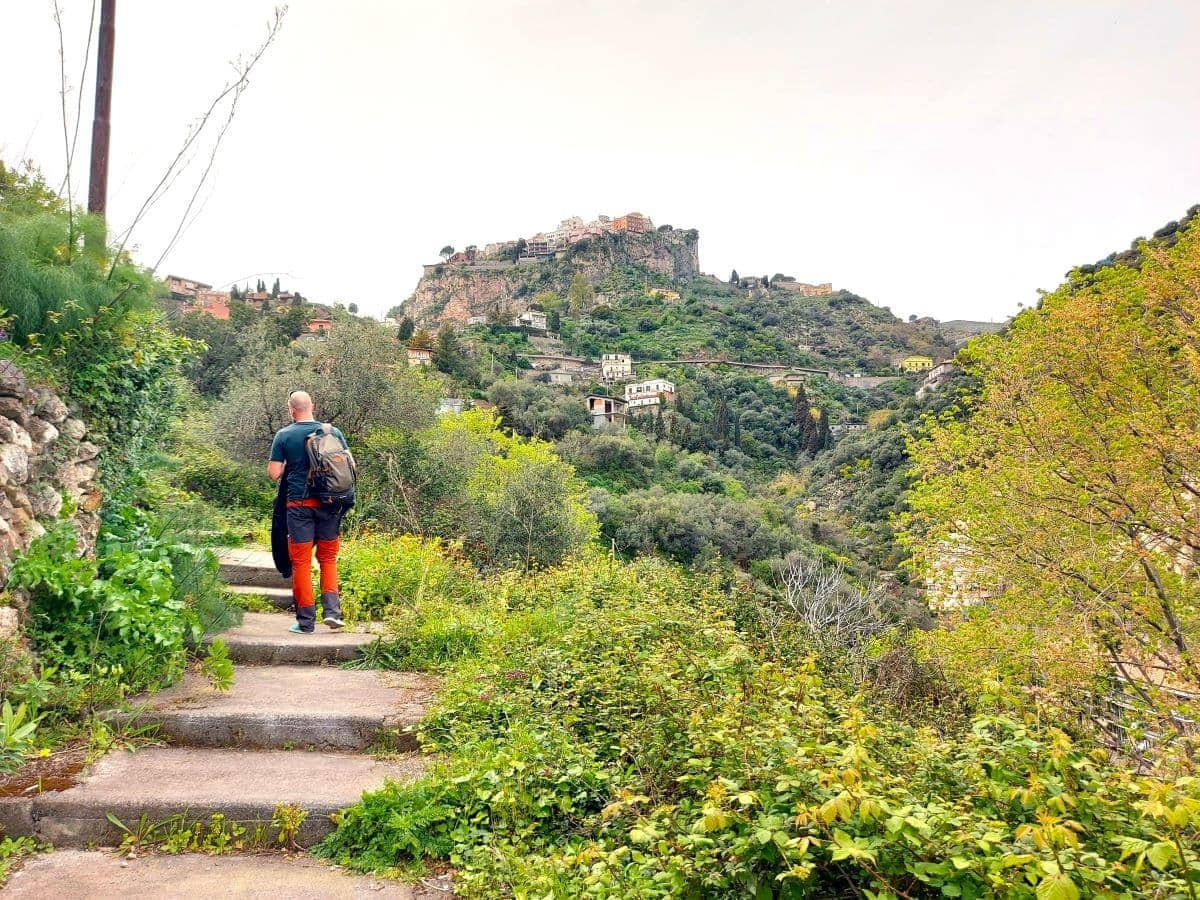 Jan in bright orange pants ascends a stone staircase on the Castelmola Taormina Walk, with the verdant hillside opening up. In a distant view of the historic town of Castelmola perched on a mountain, inviting adventure among the rich Sicilian landscape.