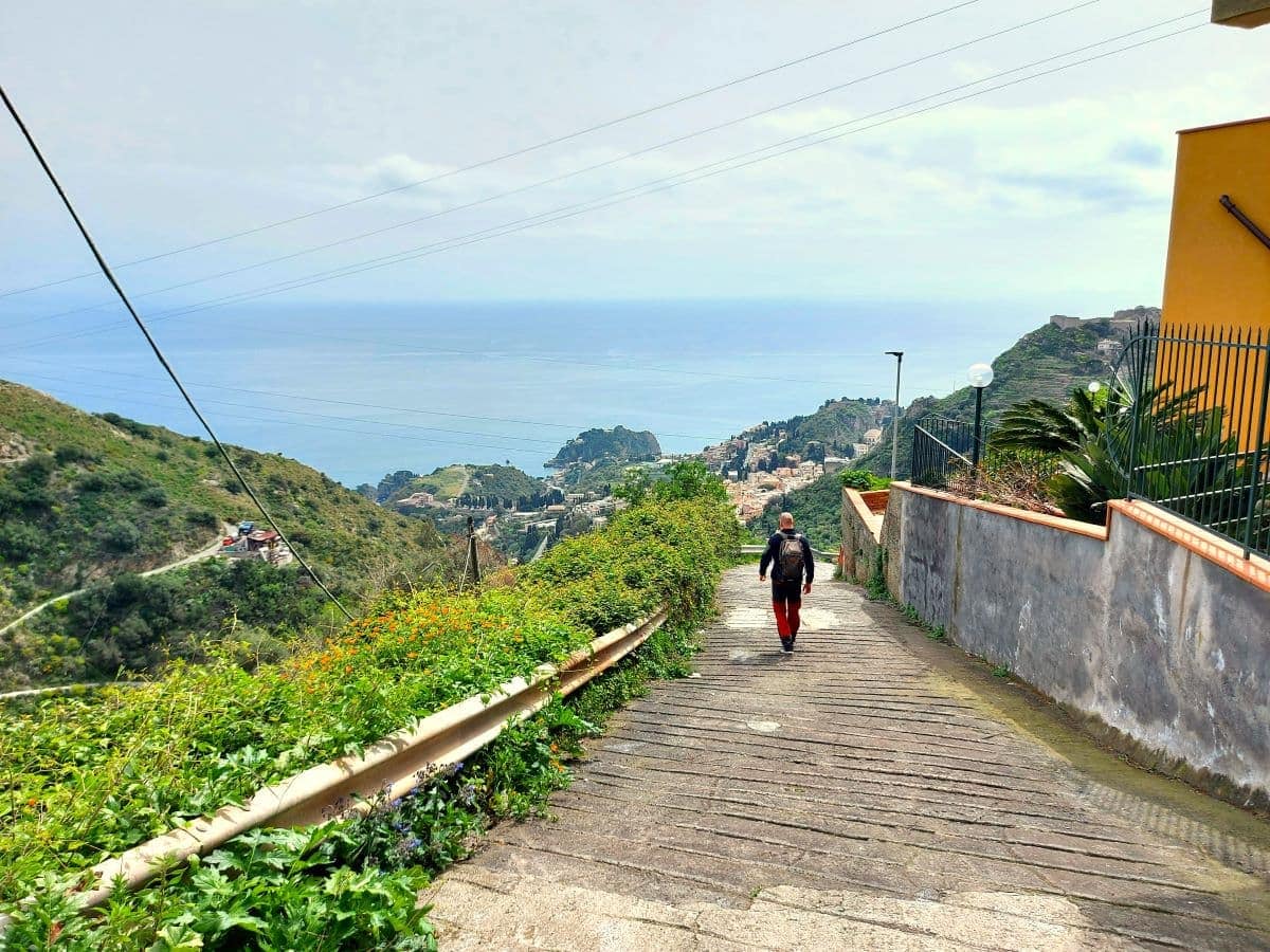 Jan, in a red jacket, treads the cobblestoned Castelmola Taormina Walk, surrounded by lush greenery and vibrant landscape with views of winding roads and the picturesque town of Taormina in the distance.