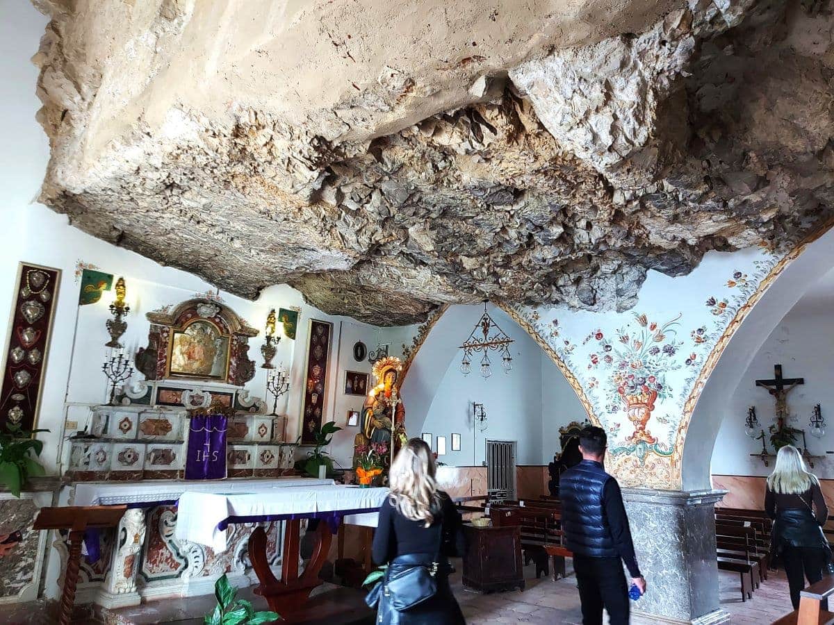 Visitors inside the Chiesa Madonna della Rocca, a unique church carved into the rock above Taormina, featuring ornate religious altarpieces, rustic stone archways, and walls adorned with vibrant fresco decorations.