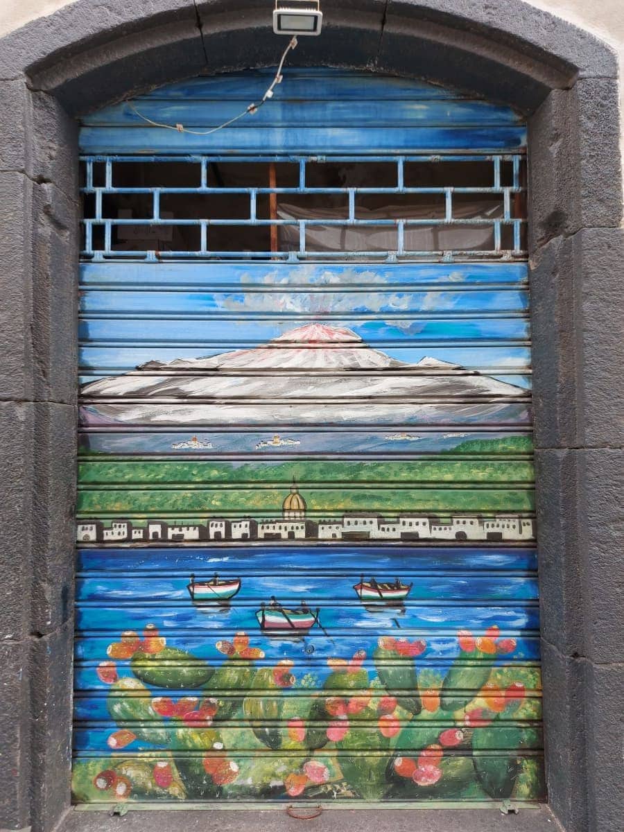 A vibrant garage door painting that depicts Mount Etna in vivid colors, complete with boats on blue waters and red prickly pears in the foreground, capturing the essence of Sicilian culture and landscape.