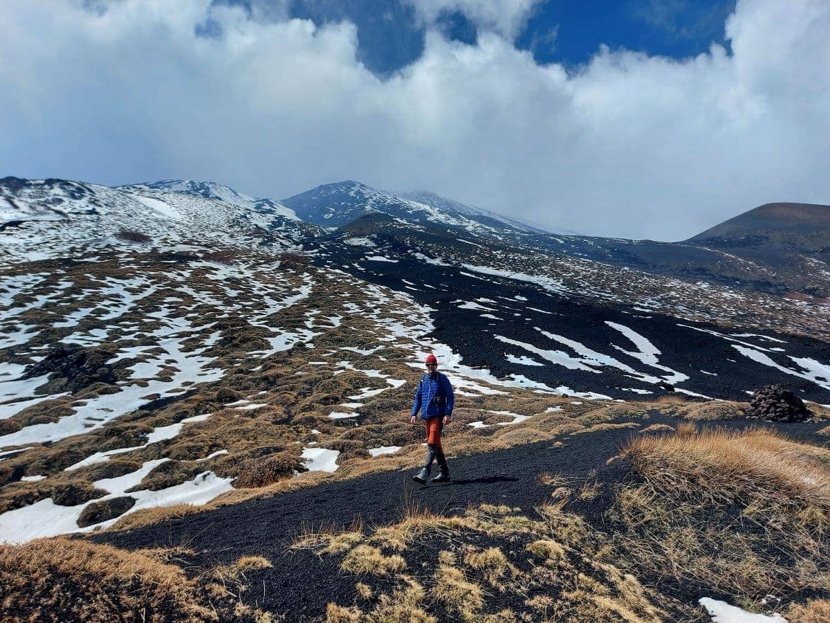 A vibrant contrast unfolds as the lone hiker Linda in bright blue and orange attire traverses the snow-streaked black volcanic terrain of Mount Etna, with the mountain's vast slopes stretching into the distance under a dynamic sky.
