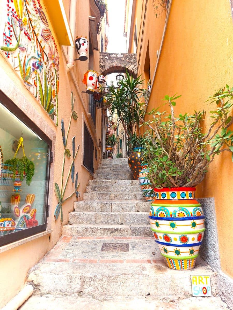 A narrow stairway in Taormina lined with vivid Sicilian ceramic pots and intricate murals, inviting visitors into the heart of the town's artistic soul, with an art shop sign adding a splash of local culture to the scene.