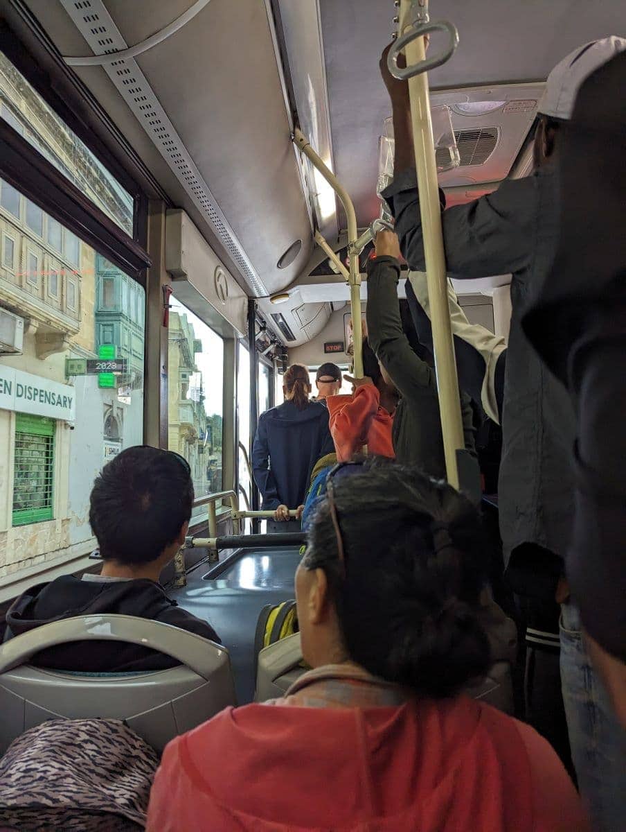 Innenraum eines überfüllten Busses in Valletta, Malta, mit stehenden und sitzenden Fahrgästen während der Fahrt. Die Ansicht fängt den alltäglichen Pendelverkehr mit einem Blick auf die Stadt durch die Busfenster ein.