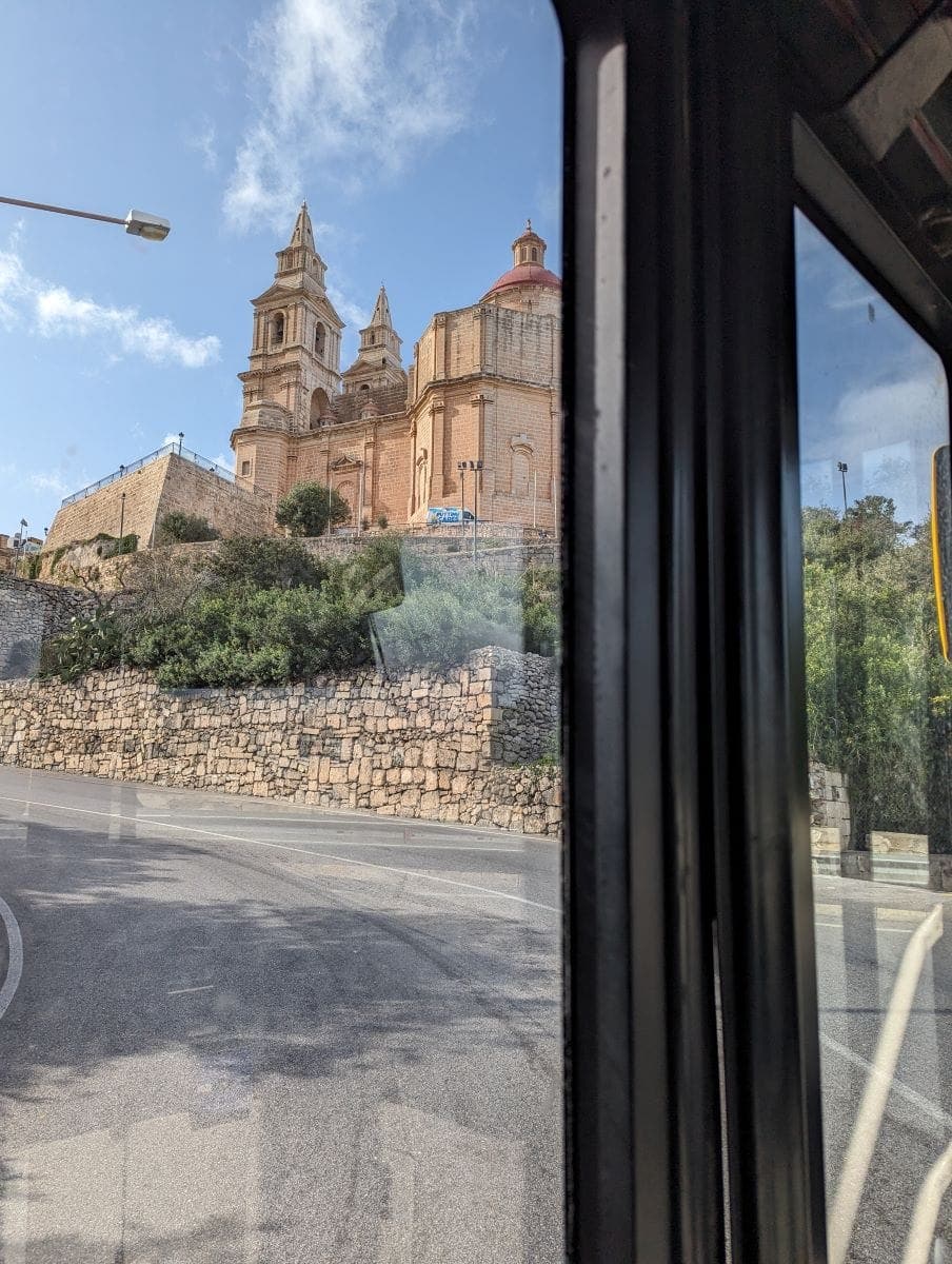 View from inside a bus on Malta, showing the baroque architecture of a church with two bell towers through the window. The reflection on the glass adds a translucent overlay to the sunlit stone structure against a blue sky