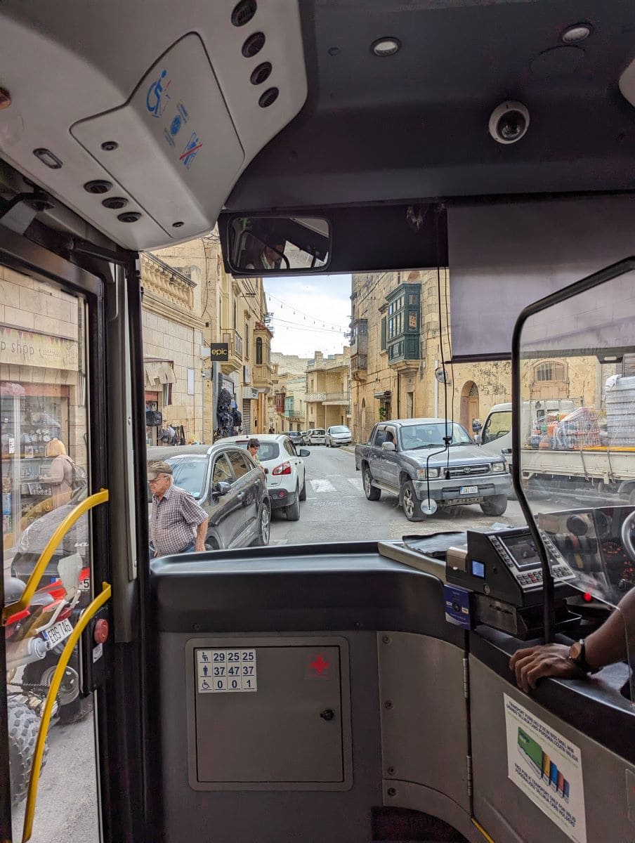 Inside view of a bus on Gozo with passengers and a driver visible. The bus is navigating through a narrow street lined with traditional Maltese buildings and parked cars, capturing the essence of daily life and transport on the island.