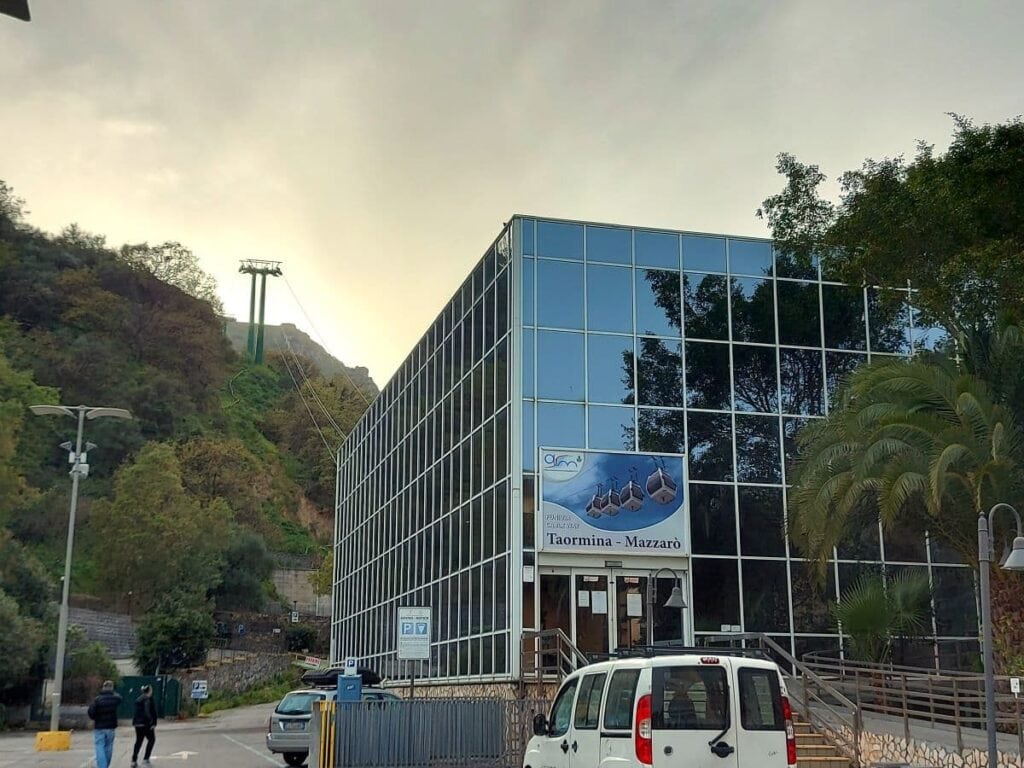 The Taormina-Mazzarò cable car station presents modern glass architecture that reflects the surrounding green hills, under a sky transitioning to dusk.