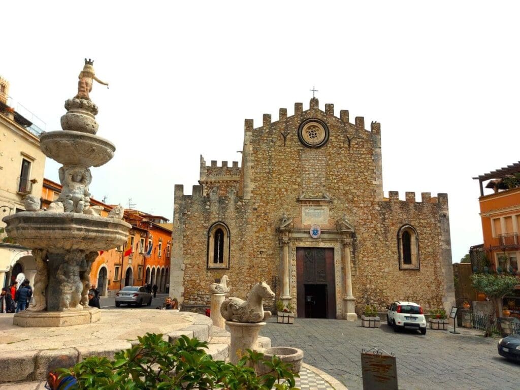 The historic Taormina Cathedral stands with solemn grace, its stone façade and crenelated battlements echoing the town's medieval past, with an ornate fountain in the foreground adding a touch of Renaissance charm to the tranquil piazza.