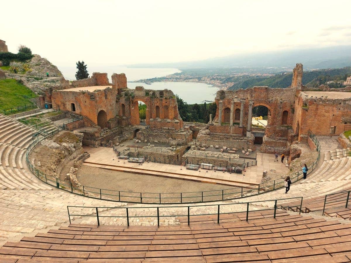 The majestic ruins of Taormina's Greek theater, set against a panoramic view of the sea, display the architectural marvels of antiquity, with its tiered seating and arches still echoing the grandeur of past spectacles.