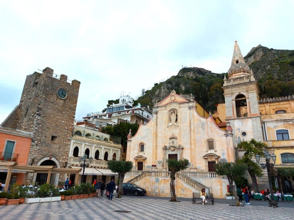 The lively Piazza IX Aprile in Taormina's old town showcases the diverse architectural heritage with a robust medieval clock tower Porta di Mezzo, the pastel-hued baroque church San Giuseppe, and a backdrop of rugged hills. All under a soft sky that promises adventure and history at every corner.