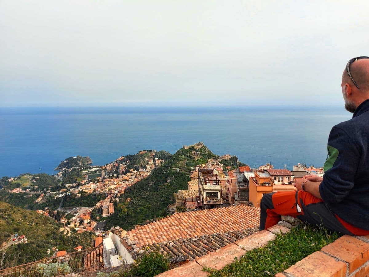 Jan takes in the expansive view from Castelmola, overlooking the terracotta rooftops of Taormina, the verdant hillside, and the azure waters of the Ionian Sea, with the picturesque Isola Bella in the distance.