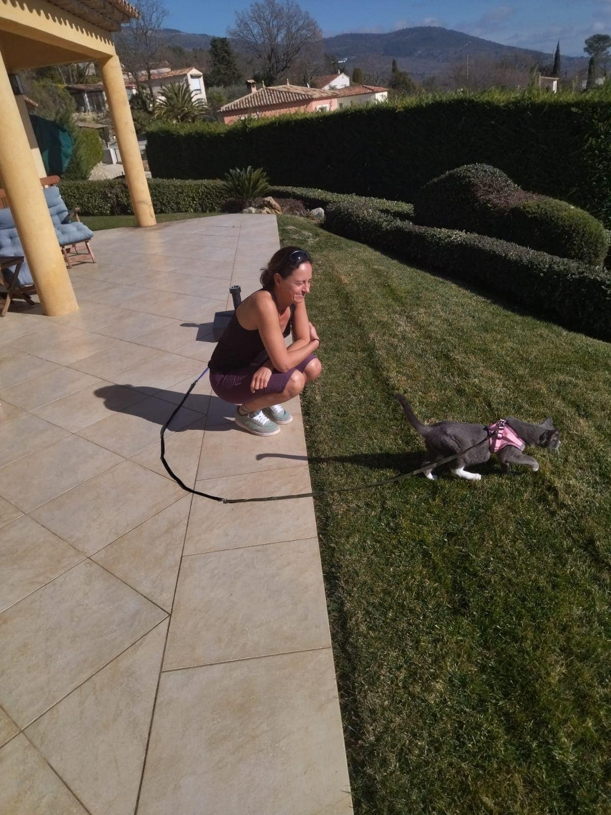Linda hockt auf einer sonnigen Terrasse und schaut einer angeleinten Katze beim Spazierengehen über den Rasen zu. Diese Szene zeigt den Alltag als Haussitterin mit Tierbetreuung in einem privaten Zuhause.