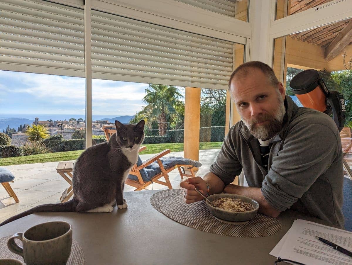 Ein Mann isst Müsli an einem Tisch mit Aussicht auf die Côte d’Azur, während eine Katze neben ihm sitzt. Das Bild zeigt, wie eng Haussitter in das Alltagsleben ihrer tierischen Mitbewohner eingebunden sind.