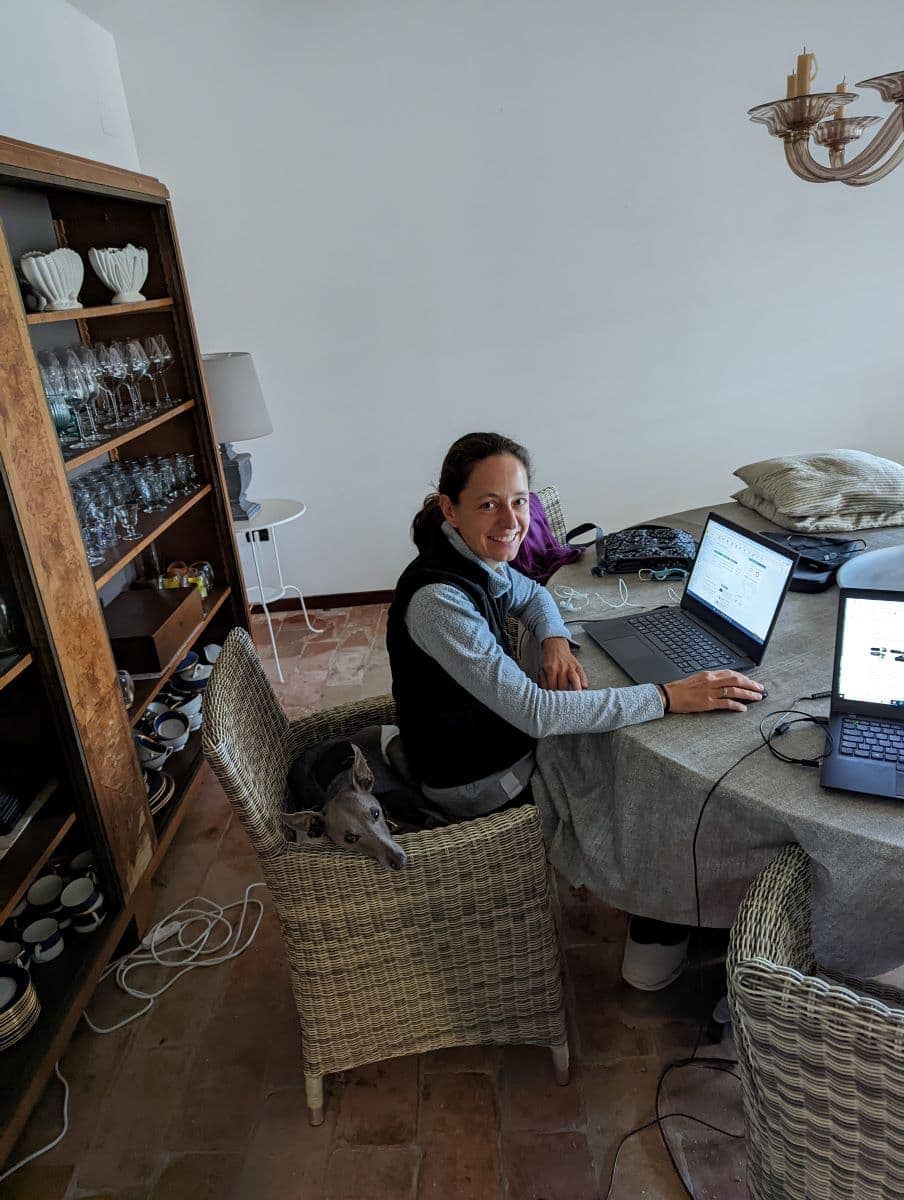 Linda arbeitet an einem Esstisch mit zwei Laptops, während ein kleiner Hund auf ihrem Stuhl liegt. Haussitter werden bietet die Möglichkeit, ortsunabhängig zu arbeiten und Tiere gleichzeitig liebevoll zu betreuen.