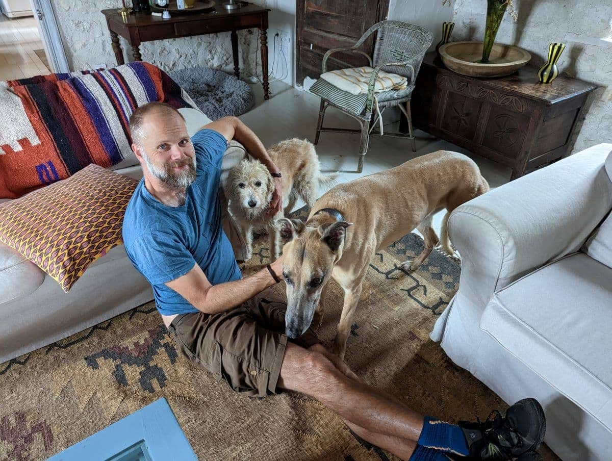 Ein Mann sitzt lächelnd auf dem Boden eines Wohnzimmers und wird von zwei Hunden umgeben, die seine Nähe suchen. Wer Haussitter werden möchte, sollte Freude an tierischer Gesellschaft und Fürsorge haben.