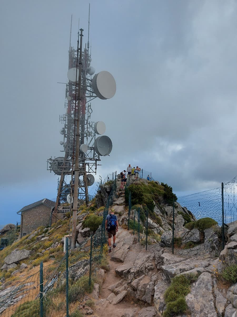 Ein Wanderer nähert sich dem Gipfel des Monte Capanne auf einem felsigen Pfad, während eine große Antenne mit zahlreichen Satellitenschüsseln den höchsten Punkt markiert. Um den Gipfel sind mehrere Besucher hinter einem Maschendrahtzaun zu sehen, während sich die Szenerie in dichten Wolken hüllt und eine mystische Atmosphäre schafft.