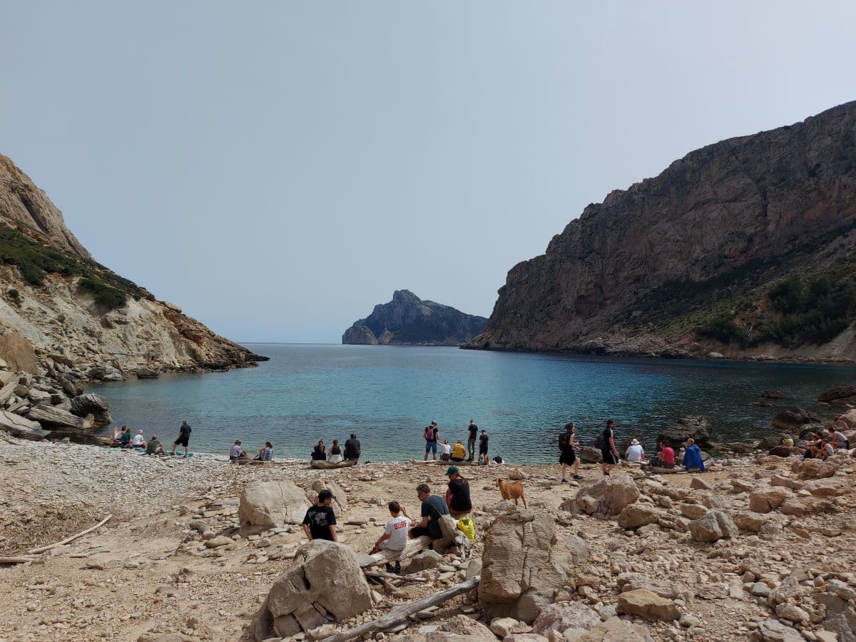 Linda and other tourists relax on the rocky beach of Cala Boquer, surrounded by towering cliffs and crystal-clear blue water, under a hazy sky.