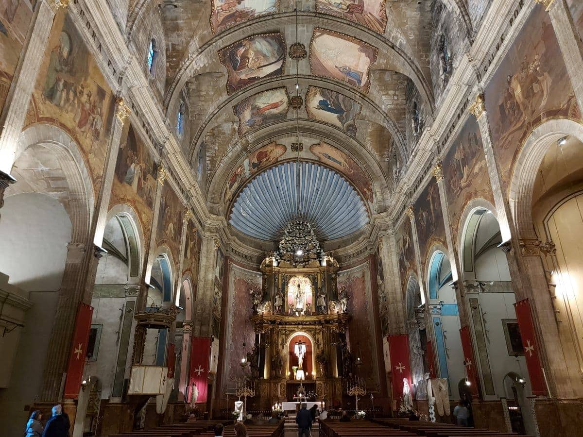 The interior of the large church in Pollensa with an ornate golden altarpiece, elaborate frescoes on the vaulted ceilings and flags on the walls, which Linda and other visitors admire.