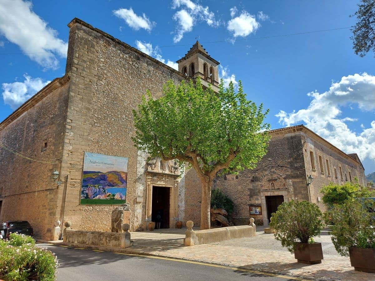 View of a stone church in Pollensa with a bell tower and a living painting on the wall, surrounded by green trees and a cloudy sky.
