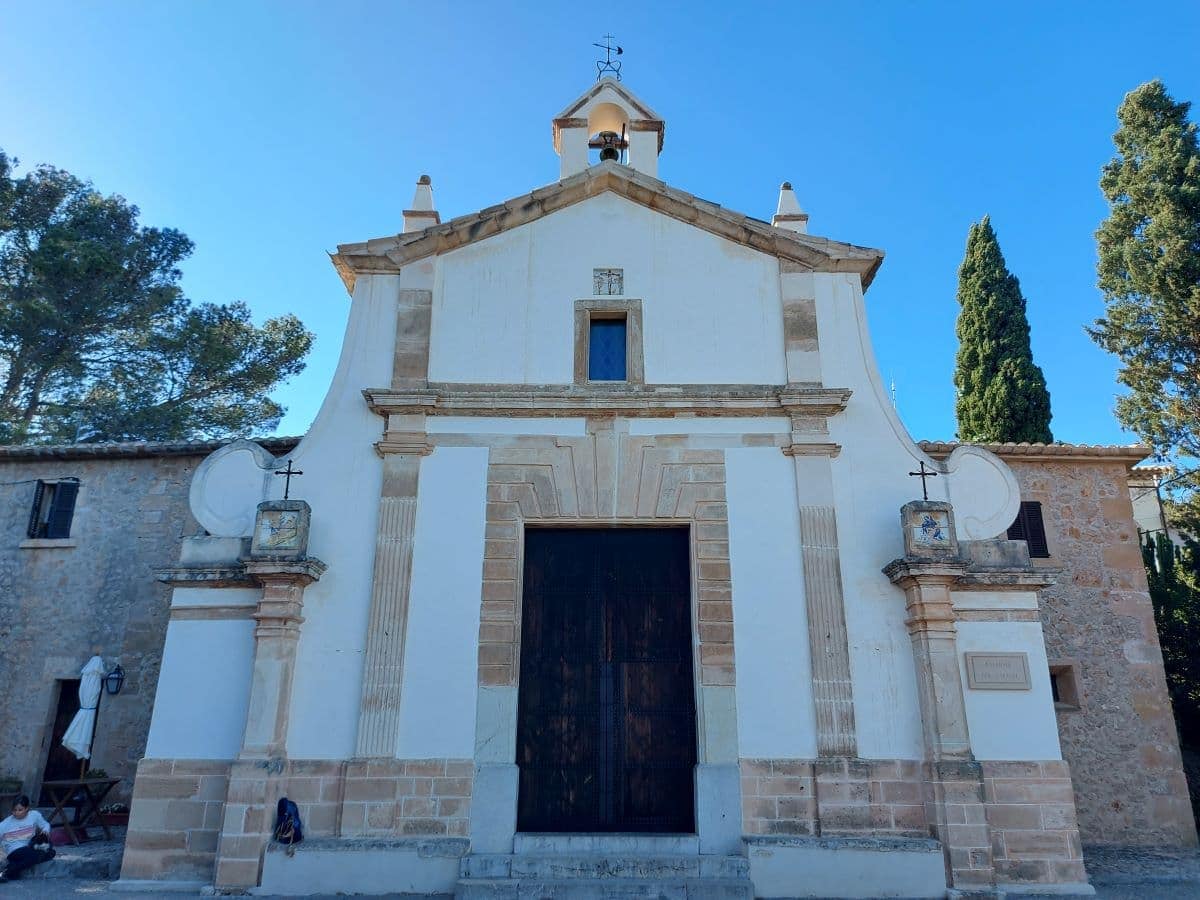 The façade of a small white church on the Calvary in Pollensa, with classic architectural details, framed by lush trees and a clear blue sky, creates a peaceful and historic ambience.