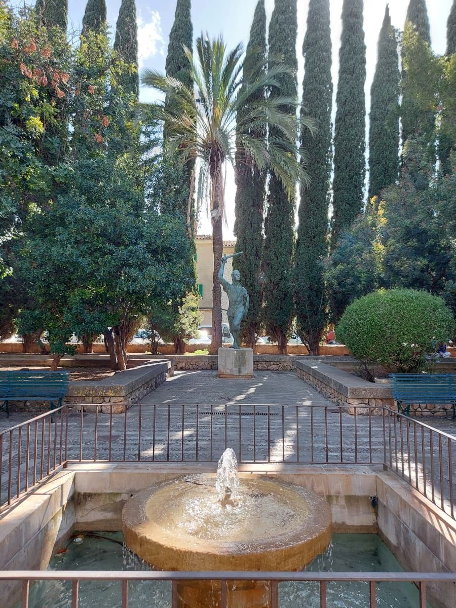 Tranquil park scene in Pollença Mallorca: small, bubbling fountain in the foreground, a statue framed by tall palm trees and cypresses, offering an empty green space for relaxation.