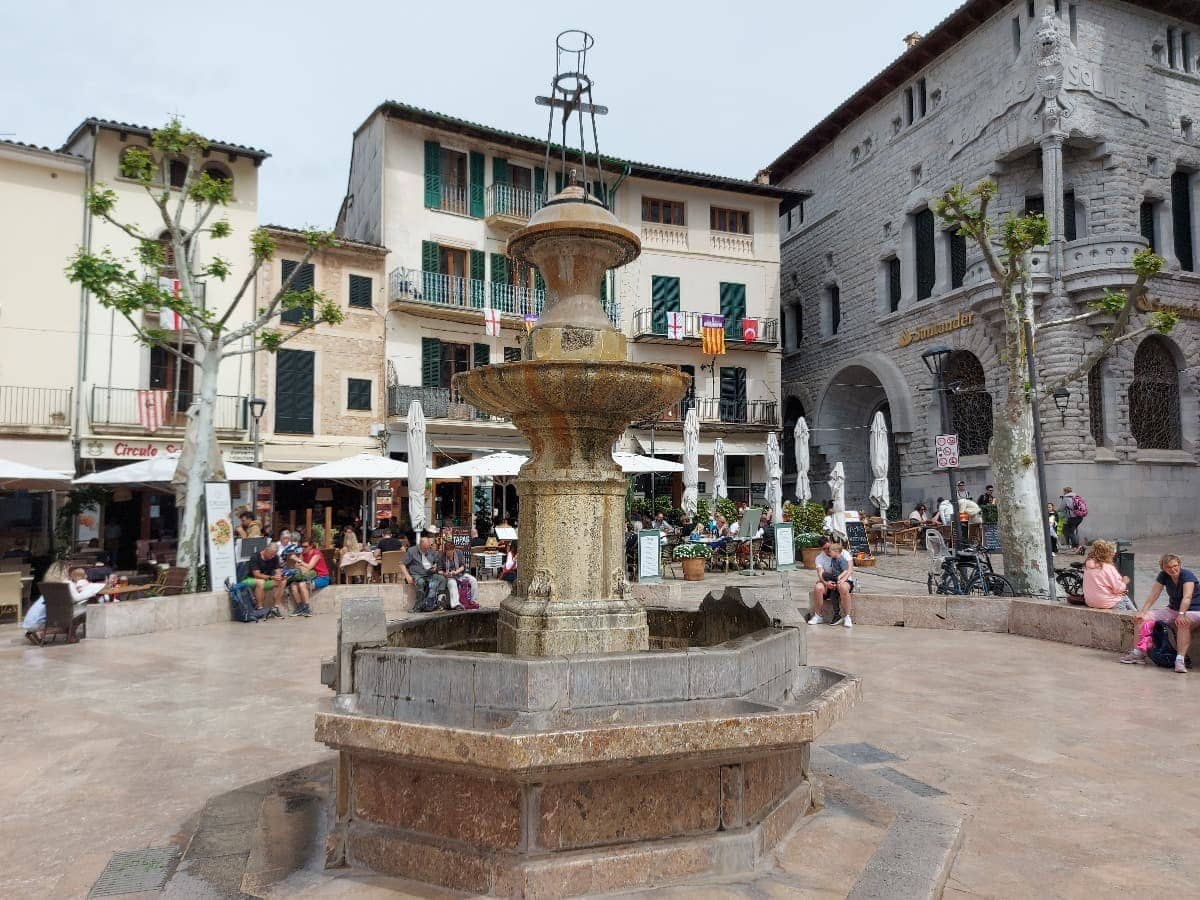 Ein zentraler Brunnen auf der Plaza de la Constitución in Sóller, umgeben von historischen Gebäuden mit Cafés und Restaurants, in denen Menschen unter Sonnenschirmen sitzen und die Atmosphäre genießen. Die Fassade der Banco de Sóller, ein markantes, steinernes Gebäude, ist rechts im Bild zu sehen, während auf der linken Seite weitere traditionelle mallorquinische Häuser die Szene prägen.