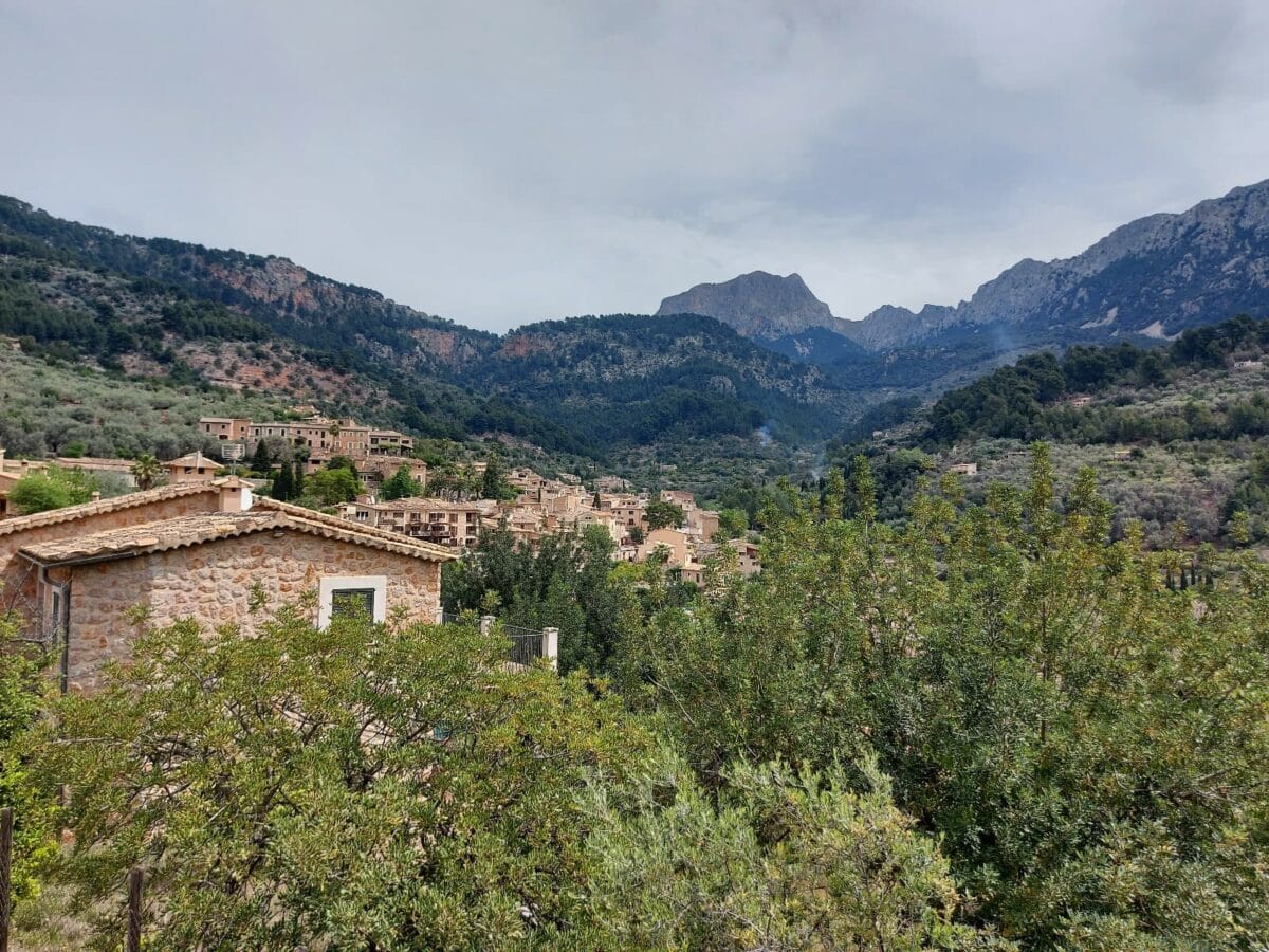 Ein malerisches Dorf in den Bergen Mallorcas, mit traditionellen Steinhäusern, die sich harmonisch in die grüne, von Olivenbäumen und üppiger Vegetation geprägte Landschaft einfügen. Im Hintergrund erheben sich die dramatischen Berge der Serra de Tramuntana, die mit ihren felsigen Gipfeln die Szenerie dominieren. Die ruhige Atmosphäre und das mediterrane Flair spiegeln die Schönheit dieser ländlichen Region wider.