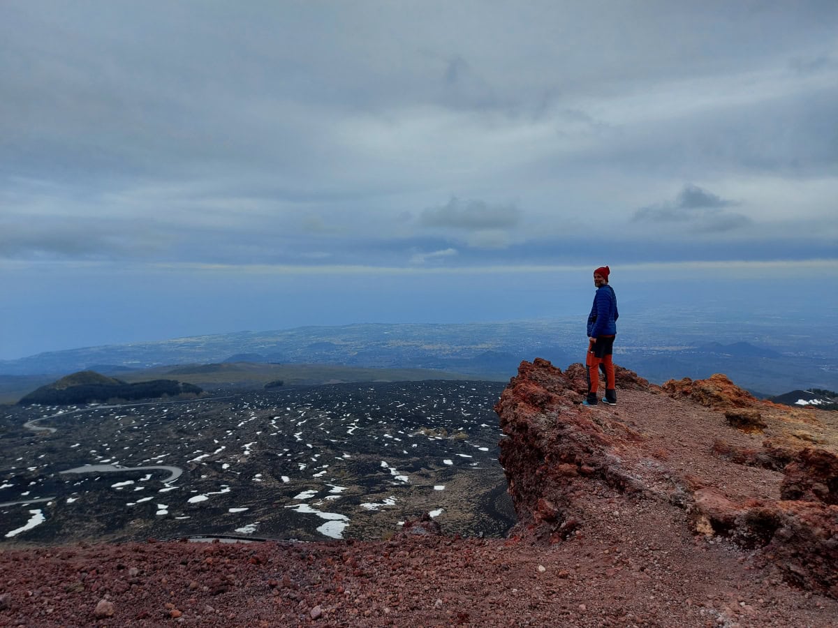 Eine Person steht auf einem rötlich-braunen Felsvorsprung nahe der Silvestri-Krater am Ätna, mit Blick über die weite Vulkanlandschaft, die von schwarzen Lavafeldern und vereinzelten Schneeflecken geprägt ist. Im Hintergrund erstrecken sich die Ebenen Siziliens bis hin zur Küste und dem Mittelmeer, sichtbar unter einem bewölkten Himmel. Die karge, dramatische Landschaft und der Kontrast zwischen den dunklen Lavasteinen und dem fernen Blau des Meeres vermitteln die majestätische Weite und Höhe des Vulkans.