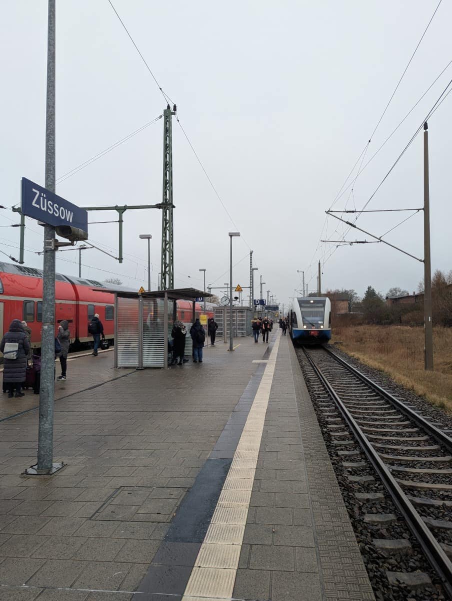Der Bahnhof Züssow mit Reisenden auf dem Bahnsteig, rechts kommt ein UBB-Zug an, links steht ein Regionalzug der Deutschen Bahn. Wenn Sie sich fragen, wie Sie nach Usedom kommen, ist Züssow ein wichtiger Umsteigepunkt - hier können Sie von einem Fernzug der Deutschen Bahn auf die UBB umsteigen, die Sie direkt auf die Insel bringt.