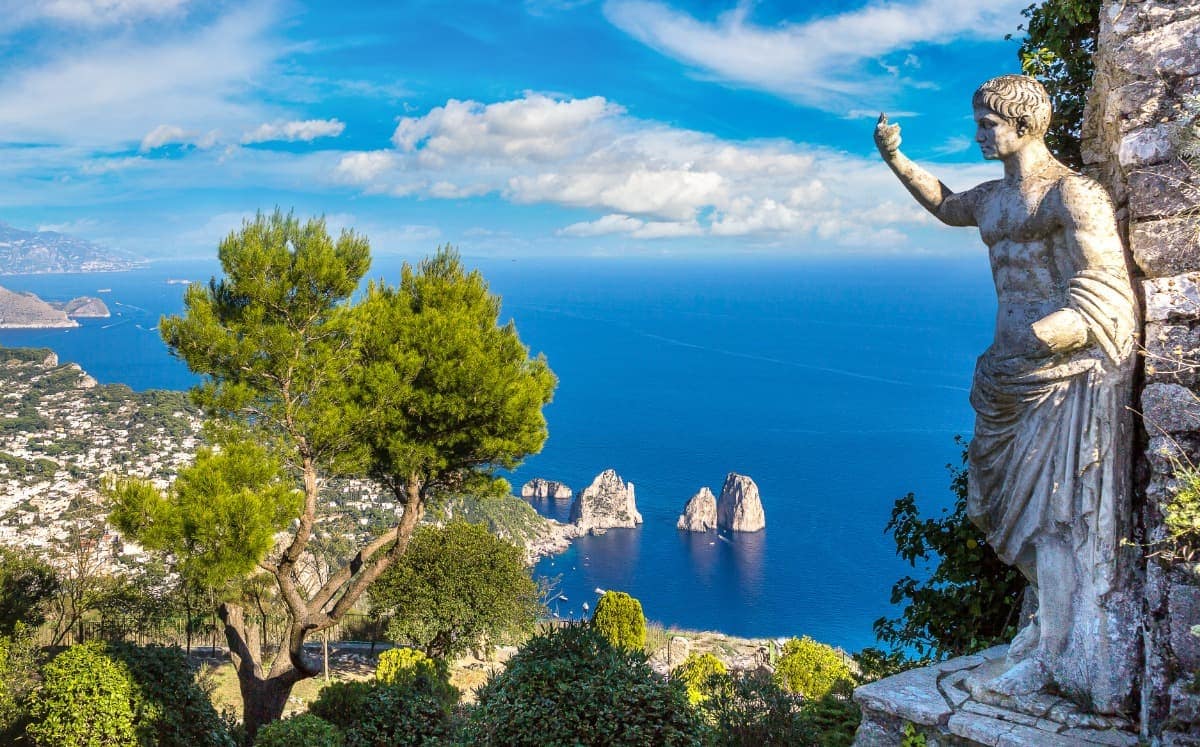 Atemberaubender Blick über die italienischen Mittelmeerinsel Capri mit einer antiken Statue im Vordergrund, die den blauen Himmel und das Meer überblickt. Umgeben von grüner Vegetation und einem majestätischen Baum sind im Hintergrund die berühmten Faraglioni-Felsen im türkisfarbenen Wasser zu sehen. Die malerische Landschaft unterstreicht die Schönheit der italienischen Küste.