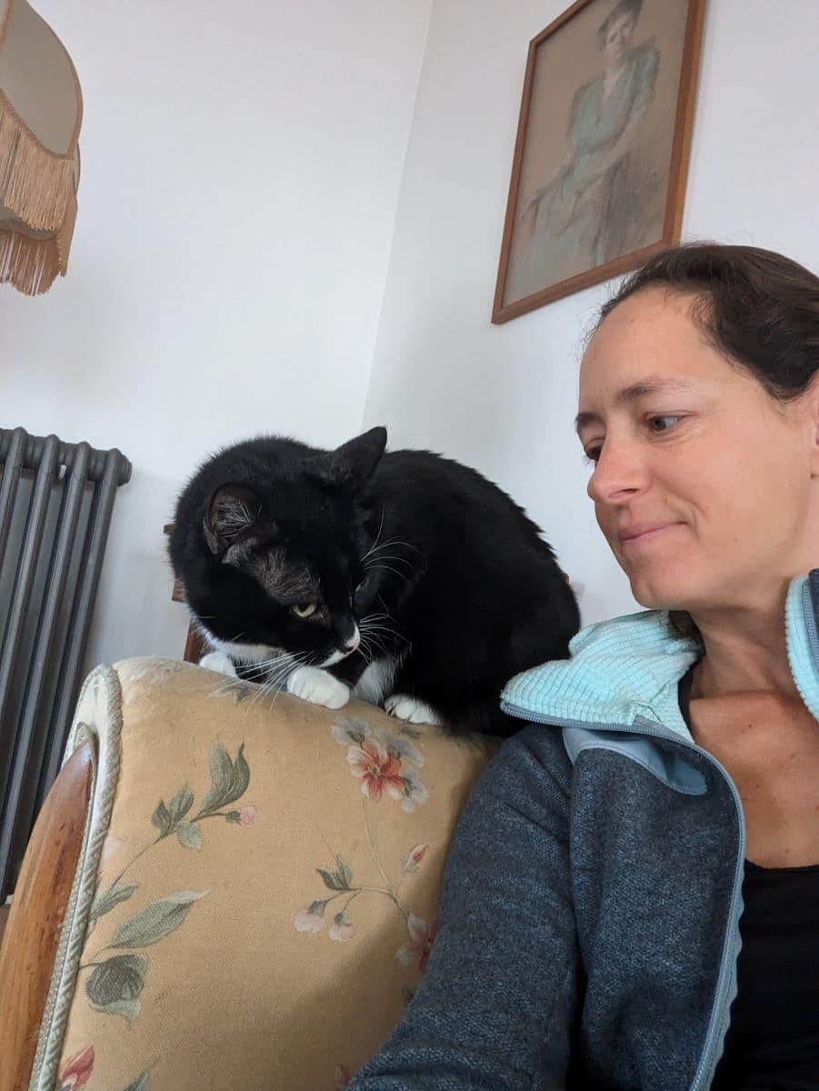 Linda sitzt in einem gemütlich eingerichteten Raum neben einer schwarz-weißen Katze auf der Sessellehne. Die Szene zeigt die Nähe und das Vertrauen, das zwischen Tieren und Haussittern entstehen kann.