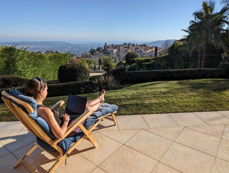Linda sitzt entspannt auf einer Sonnenliege und arbeitet am Laptop mit Blick auf ein mittelalterliches Dorf. Haussitter werden bietet die Möglichkeit, Arbeit und Reiselust in traumhafter Umgebung zu verbinden.