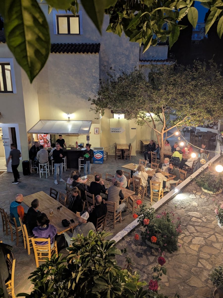 Der Innenhof des Sportclub Helen Yolanda ist abends belebt mit Gästen, die an langen Holztischen unter Bäumen zusammen sitzen – typisch gesellig im Samos Urlaub.