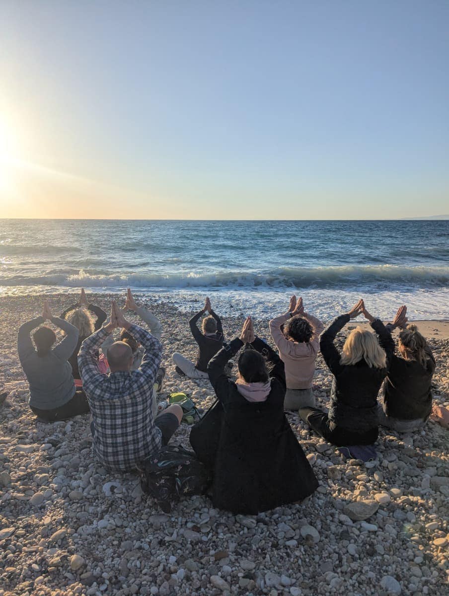 Teilnehmer:innen einer Yoga-Gruppe sitzen am Kiesstrand von Samos im Schneidersitz und meditieren beim Sonnenuntergang – ein Highlight im Urlaub auf Samos mit Froschreisen.